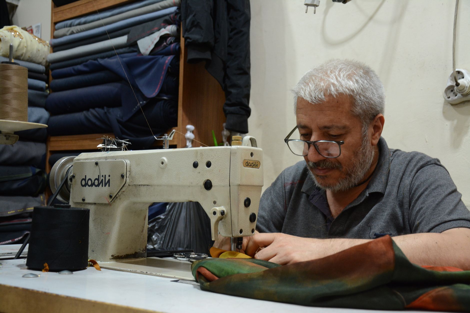 A senior tailor skillfully operates a sewing machine, surrounded by fabric in a workshop setting.
